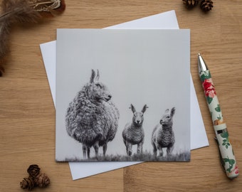 May include: A black and white greeting card featuring a sheep and two lambs standing in a field. The card is on a wooden surface with a white envelope, a pen with a floral design, pine cones, and a feather.