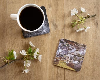 May include: A white coffee mug filled with black coffee sits on a square coaster with a marble design. A second coaster features a landscape with a goat. Cherry blossoms are arranged around the coasters on a wood surface.