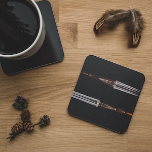 May include: A black square coaster with a photo of two silver shotguns on a black background. The coaster is on a wooden surface with a cup of coffee and a feather.