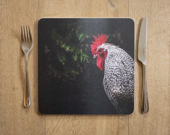 May include: Square placemat featuring a close-up of a rooster with black and white speckled feathers, a red comb and wattle, and a dark background. A fork and knife are on either side of the placemat, set on a wooden surface.