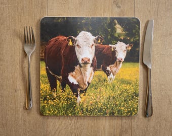 May include: A square placemat featuring a pastoral scene with two brown and white cows standing in a field of yellow flowers. A fork and knife are placed on a wooden surface next to the placemat.