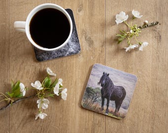 May include: A white coffee mug filled with black coffee sits on a coaster with a marble design. A second coaster features a black horse in a landscape. White flowers and greenery are arranged on a wooden surface.