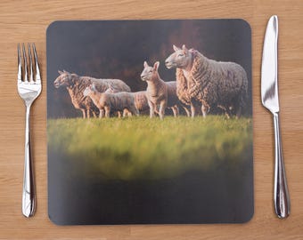 May include: A square placemat featuring a pastoral scene of sheep grazing in a green field under a dark sky. A fork and knife are on either side of the placemat, set on a wooden table.