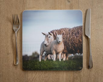 May include: Square placemat featuring a pastoral scene of two sheep standing in a grassy field, with a larger sheep behind them. A fork and knife are on either side of the placemat, set on a wooden surface.