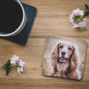 Può includere: Un sottobicchiere quadrato con un ritratto di un cane Cocker Spaniel. Il cane ha il pelo marrone chiaro e la faccia bianca. Una tazza di caffè è su un sottobicchiere nero. Gli oggetti sono su una superficie di legno con fiori.