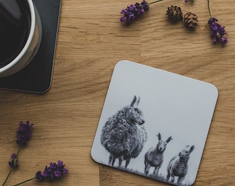 May include: A white ceramic coaster with a black and white illustration of three sheep. The sheep are standing in a line, with the largest sheep in the front and the smallest sheep in the back.