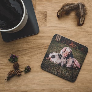 May include: A square coaster with a photograph of two piglets cuddling in a grassy field. The piglets are pink with black spots.