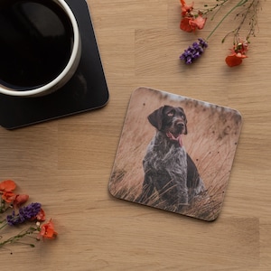May include: A brown and grey dog with a long tongue sits in a field of tall grass. The dog is looking to the right of the image. The dog is on a square coaster.