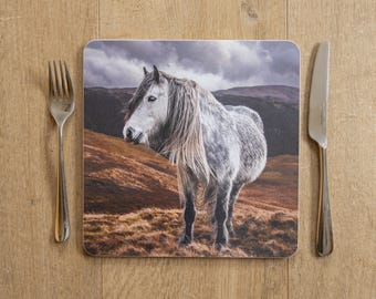 May include: Square coaster featuring a majestic white horse with a long mane, set against a backdrop of rolling hills and a cloudy sky. A fork and knife are placed on either side of the coaster on a wooden surface.