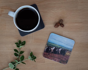 May include: A white mug filled with dark coffee sits on a dark gray square coaster. A second coaster displays a landscape with black and white cows. A sprig of green and white leaves and two pinecones are also on the wooden surface.