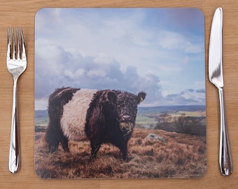 May include: Square placemat featuring a black and white cow in a field under a cloudy sky. A silver fork and knife are on either side of the placemat, set on a wooden surface. The placemat is a decorative item for the dining table.