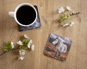 May include: A white coffee mug filled with black coffee sits on a marble-patterned coaster. A coaster with a horse image is next to a sprig of white flowers. The items are arranged on a wooden surface.