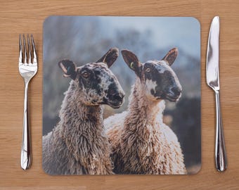 May include: A square placemat featuring a photographic print of two sheep with brown and white faces and fluffy, light brown wool. A silver fork and knife are placed on a wooden surface, framing the placemat.