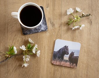May include: A white coffee mug filled with dark liquid sits on a gray and white marble-patterned coaster. A second coaster features two black horses in a field. White flowers and greenery are arranged around the items.