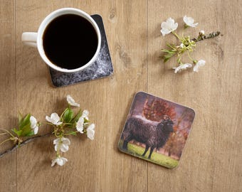 May include: A white mug filled with black coffee sits on a coaster with a marble design. A second coaster features a black sheep on a green field. White flowers and branches are arranged on a wooden surface.