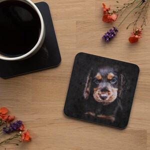 May include: Square black coaster featuring a close-up of a Dachshund puppy with brown and black fur and blue eyes. A matching coaster and a cup of coffee are visible. The background is a light wood surface.