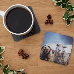 May include: A white coffee mug filled with dark liquid sits on a black square coaster. A second coaster features a photo of two sheep. The scene is set on a wooden surface with decorative greenery and pinecones.