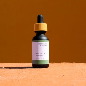 Puede incluir: Una botella de tintura negra con cuentagotas y tapón de madera. La etiqueta dice "Mullein Tincture" y "Better Ours + Garden". La botella es de 30 ml. El fondo es de color naranja.