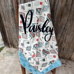 Op de afbeelding: Een wit en blauw geometrisch patroon babydeken met een turquoise nep bont rand. De deken heeft de naam "Paisley" in het zwart op de voorkant.