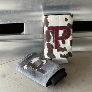 Pode incluir: Dois porta-copos de tecido com a letra "P" bordada. O porta-copos &agrave; esquerda &eacute; cinza com um "P" de estampa de vaca marrom e branco. O porta-copos &agrave; direita &eacute; branco com estampa de vaca marrom e branco e um "P" bord&ocirc;.