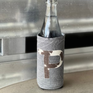 Pode incluir: Um porta-garrafas de tecido cinza com uma letra "P" de estampa de vaca branca e marrom na frente. O porta-garrafas est&aacute; em uma garrafa de vidro.