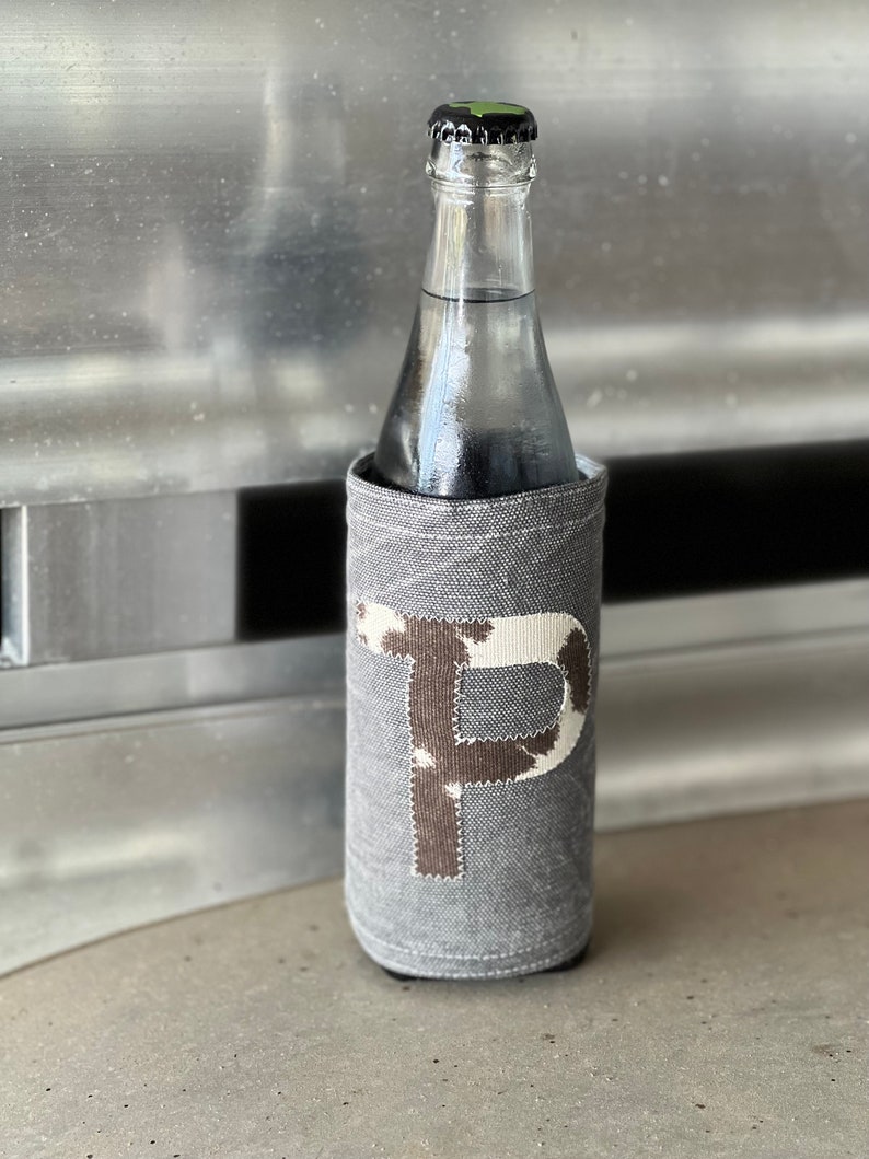 Pode incluir: Um porta-garrafas de tecido cinza com uma letra "P" de estampa de vaca branca e marrom na frente. O porta-garrafas est&aacute; em uma garrafa de vidro.
