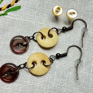 Puede incluir: Un par de pendientes hechos con dos tipos diferentes de botones. Un pendiente tiene un botón marrón con un brillo nacarado y un botón blanco con dos agujeros. El otro pendiente tiene un botón blanco con dos agujeros y un botón marrón con un brillo nacarado.