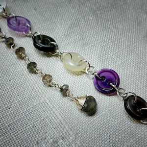 May include: A silver chain bracelet with alternating purple, white, and gray buttons and dark gray gemstone beads.