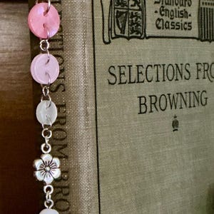 May include: A handmade bookmark with a silver-tone clip and a dangling charm. The charm features pink buttons, white beads, and a silver flower. The bookmark is placed in a book titled "Selections From Browning."