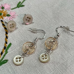 Może przedstawiać: Para kolczyków z przezroczystą szklaną koralikiem i białym guzikiem. Kolczyki są wykonane ze srebrnego metalu.