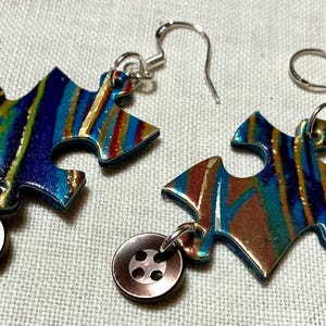 Puede incluir: Un par de pendientes únicos con forma de piezas de rompecabezas con un diseño colorido y arremolinado en azul, verde y dorado. Cada pendiente tiene un pequeño dije de botón redondo marrón. Los pendientes tienen ganchos plateados.