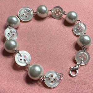 May include: A handmade bracelet featuring alternating faux pearl beads and translucent, iridescent buttons. The bracelet is connected with silver-colored jump rings and has a silver-colored clasp. The bracelet is displayed on a pink surface.