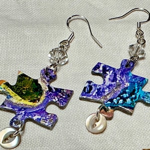 May include: A pair of unique dangle earrings. Each earring features a puzzle piece shape with a colorful abstract design in shades of blue, purple, and green. Clear beads and a small button accent the earrings, which hang from silver-tone hooks.