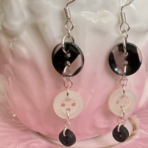 May include: A pair of dangle earrings featuring black and white buttons with silver chain links. The top button is black with white stripes, the middle button is white with four holes, and the bottom button is black.