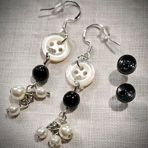 Puede incluir: Un par de pendientes con abalorios blancos con forma de botón, cuentas negras y detalles de perlas. Los pendientes cuelgan de ganchos plateados. También hay dos abalorios negros adicionales con forma de botón.