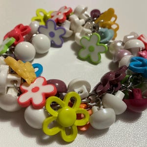 May include: A vibrant bracelet with a mix of charms: white beads, flower shapes in pink, blue, green, and yellow, and mushroom-shaped charms. The bracelet is made of metal and plastic.