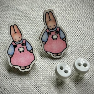 Puede incluir: Un par de pendientes de madera de conejo con vestidos rosas y un diseño de oveja blanca. Los pendientes vienen con dos respaldos de botón blancos.