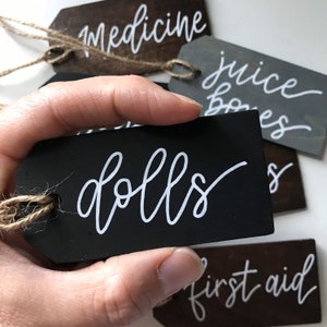 Peut inclure: &Eacute;tiquette en bois noir avec l'inscription "dolls" en &eacute;criture cursive blanche. L'&eacute;tiquette est attach&eacute;e avec de la ficelle et est tenue par une main.