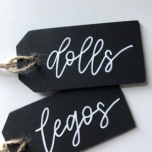 Peut inclure: Deux &eacute;tiquettes en bois noir avec de la ficelle attach&eacute;e. L'&eacute;tiquette du haut dit "dolls" en &eacute;criture cursive blanche. L'&eacute;tiquette du bas dit "legos" en &eacute;criture cursive blanche.