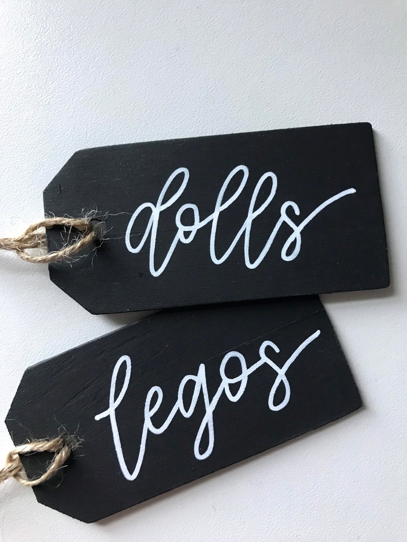 Peut inclure: Deux &eacute;tiquettes en bois noir avec de la ficelle attach&eacute;e. L'&eacute;tiquette du haut dit "dolls" en &eacute;criture cursive blanche. L'&eacute;tiquette du bas dit "legos" en &eacute;criture cursive blanche.