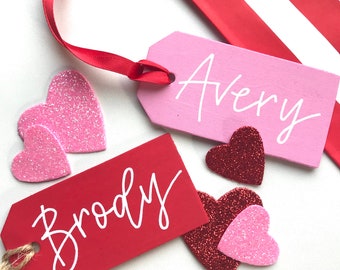 Könnte beinhalten: Zwei rosa und rote Geschenkanhänger mit den Namen Avery und Brody in weiß. Die Anhänger sind von kleinen roten und rosa Glitzerherzen umgeben.