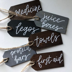 Peut inclure: Six &eacute;tiquettes en bois avec une ficelle attach&eacute;e, chacune avec une &eacute;tiquette diff&eacute;rente &eacute;crite en calligraphie blanche. Les &eacute;tiquettes indiquent "m&eacute;dicaments", "bo&icirc;tes de jus", "l&eacute;gos", "serviettes", "barres prot&eacute;in&eacute;es" et "premiers soins".