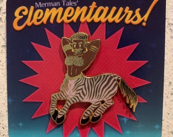 May include: Gold-colored enamel pin featuring a zebra centaur with a human torso, black hair, and a pearl necklace. The centaur is wearing a pink and white striped bikini top and has a gold-colored halo. The pin is on a blue and pink background with the text "Merman Tales' Elementours!" in gold lettering.