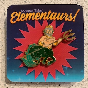 May include: A gold-colored pin featuring a cartoon centaur with a trident. The centaur has a tan torso, green tail, and a crown. The pin is set against a red starburst on a blue background with the text "Merman Tales' Elementaurs!"