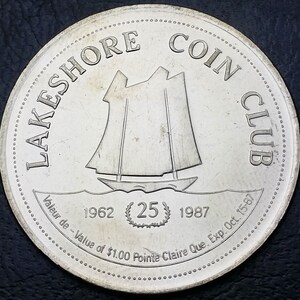 May include: A silver coin from the Lakeshore Coin Club, featuring a sailboat design. The coin has the text "LAKESHORE COIN CLUB" around the edge, with the years 1962 and 1987. The text "Valeur de - Value of $1.00 Pointe Claire Que. Exp. Oct. 15-87" is also present.