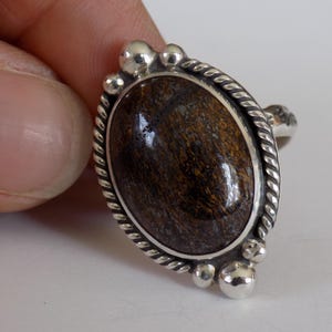 May include: A silver ring with an oval, dark brown stone. The stone is set in a silver bezel with a twisted rope design around the edge. The ring has small silver ball accents.