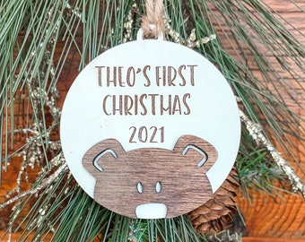 May include: A round, white Christmas ornament with the text "THEO'S FIRST CHRISTMAS 2021" in brown lettering. A wooden bear face is at the bottom. The ornament hangs from a twine string, set against a backdrop of pine needles and a wooden surface.