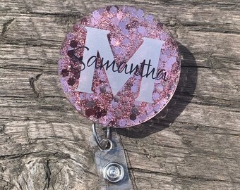 May include: A pink glitter badge holder with a white letter "M" and the name "Samantha" written in black. The badge holder is attached to a clear retractable badge reel.