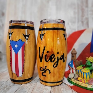 May include: Two barrel-shaped tumblers with a wood grain design. One features the Puerto Rican flag, the other has the word "Vieja" in black script and a stylized figure. Both have black bands and clear lids.