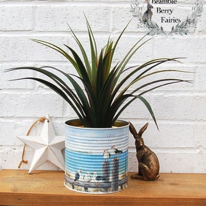 May include: A white metal tin can with a seagull design is filled with a green plant. The can is sitting on a wooden surface with a white star and a brown rabbit figurine.